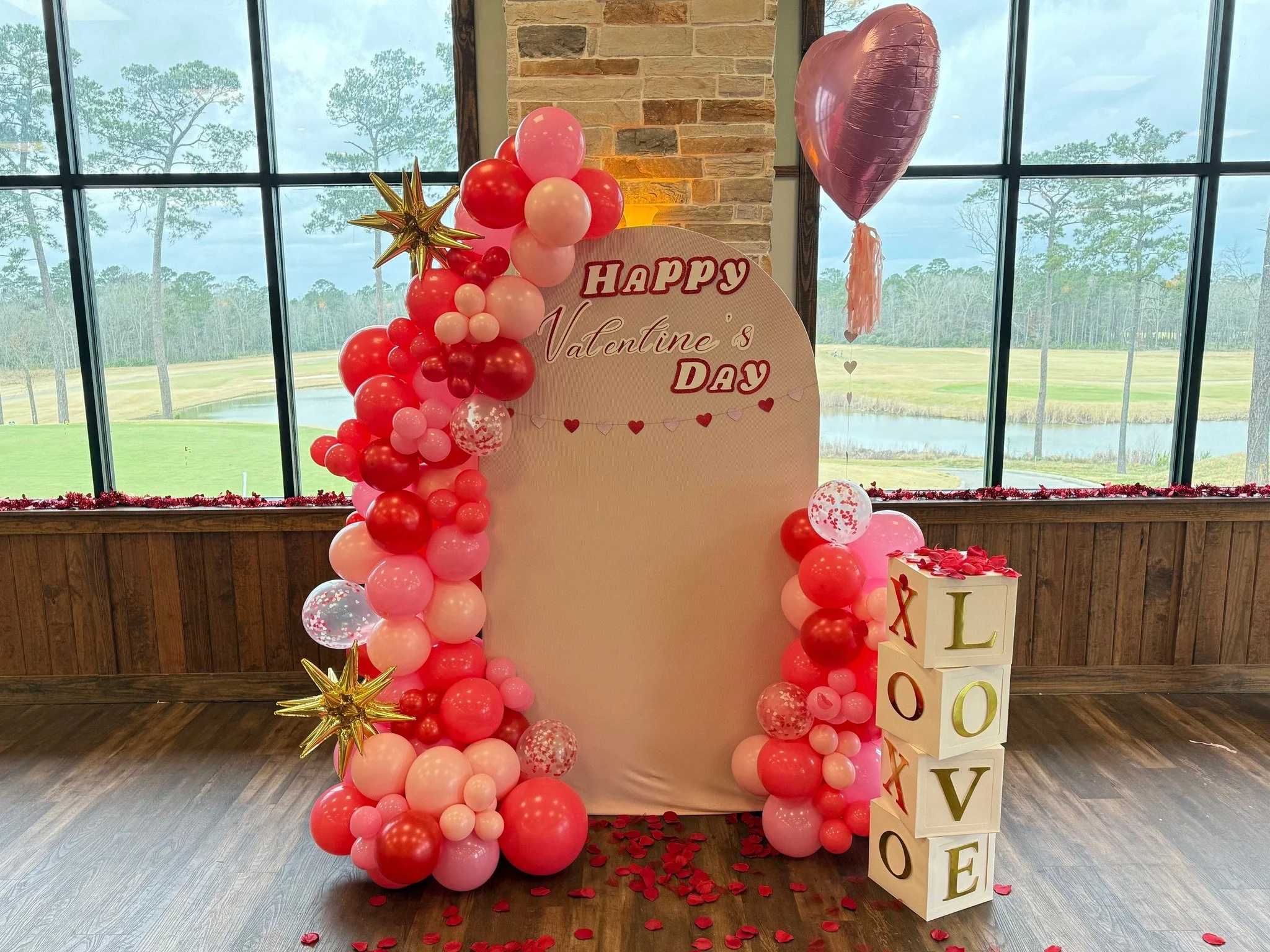 Valentine's Day celebration setup with pink and red balloons, gold star balloons, a heart-shaped pink balloon, a white backdrop with Valentine's Day message, decorative blocks spelling love, and a scenic window view.