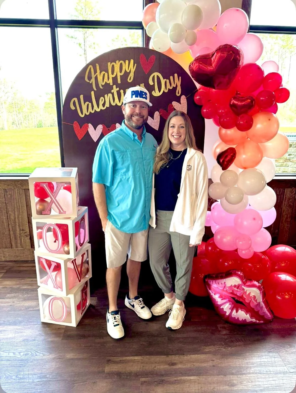 A man and woman standing together in front of a Valentine's Day decoration, with a 'Happy Valentine's Day' sign and a balloon arrangement with red, pink, and white balloons, including heart-shaped balloons and lips-shaped balloons, inside a room with