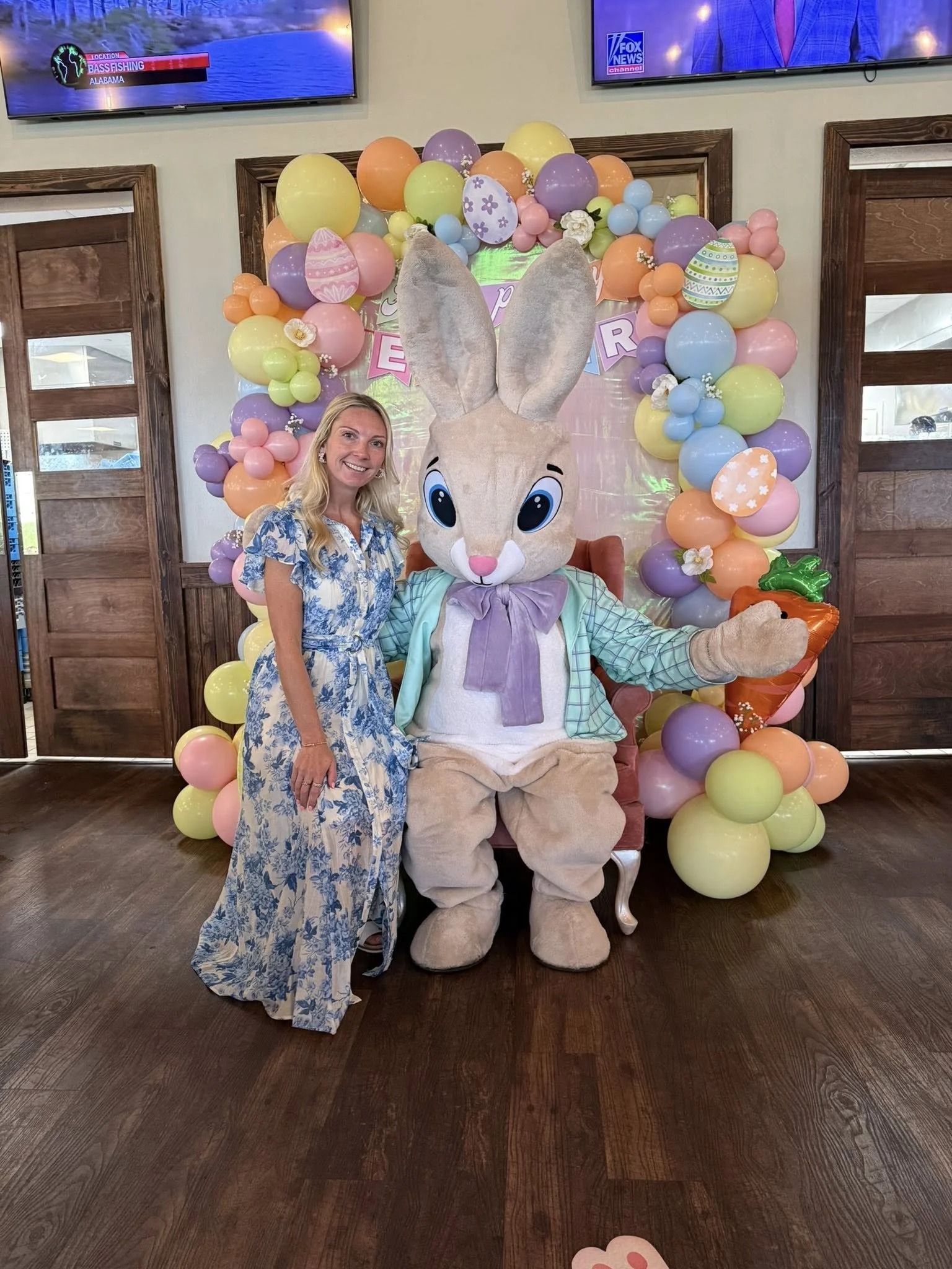 A woman in a blue floral dress standing next to a person in a large Easter Bunny costume, who is seated on a pink chair, with a colorful balloon arch behind them. The background is decorated for Easter with pastel-colored balloons and flowers.