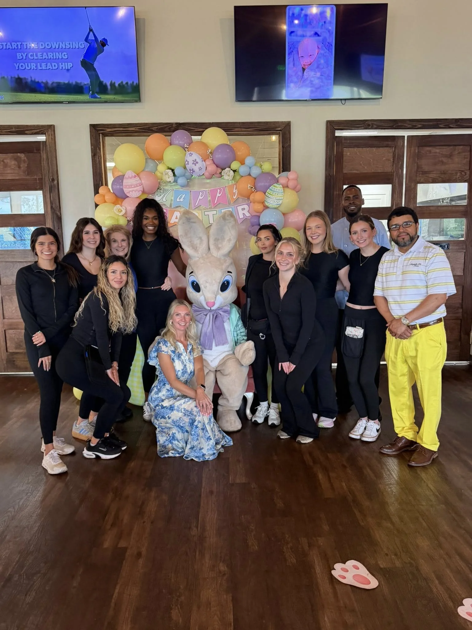 A group of people gathered around a person in a large rabbit costume at an Easter celebration. The background is decorated with pastel-colored balloons, a banner that says 'Happy Easter,' and bunny-shaped decorations on the floor. The setting appears
