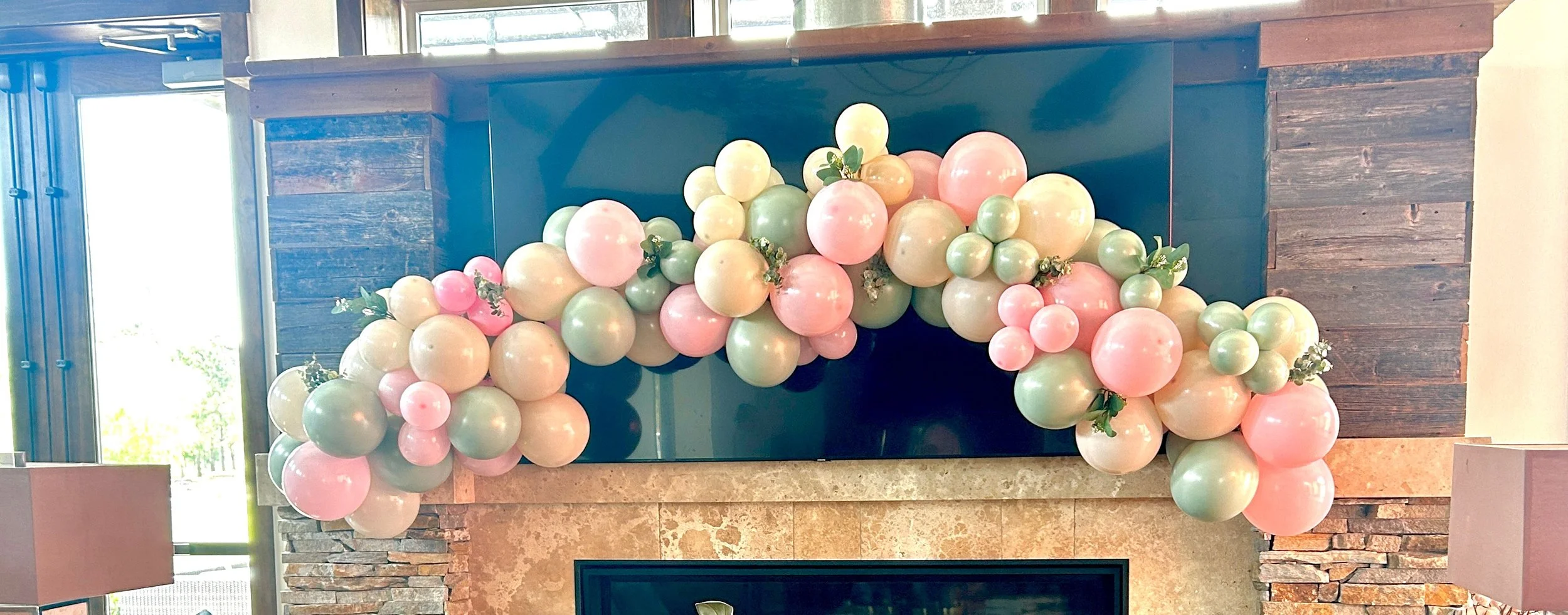 Decorative balloon arrangement with pastel pink, green, and cream balloons around a fireplace mantel in a room with large windows and wooden wall accents.