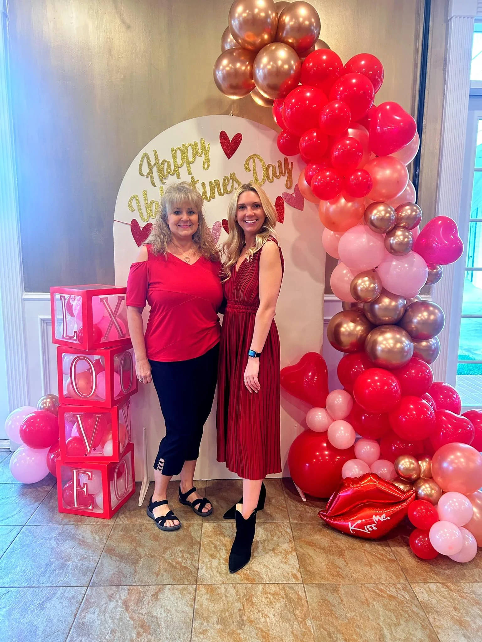 Two women standing together in front of Valentine's Day decorations, including a heart-shaped balloon arrangement, a backdrop with the words 'Happy Valentine's Day' and pink and red balloons, and a box depicting the word 'LOVE' with balloons, celebrating Valentine's Day.