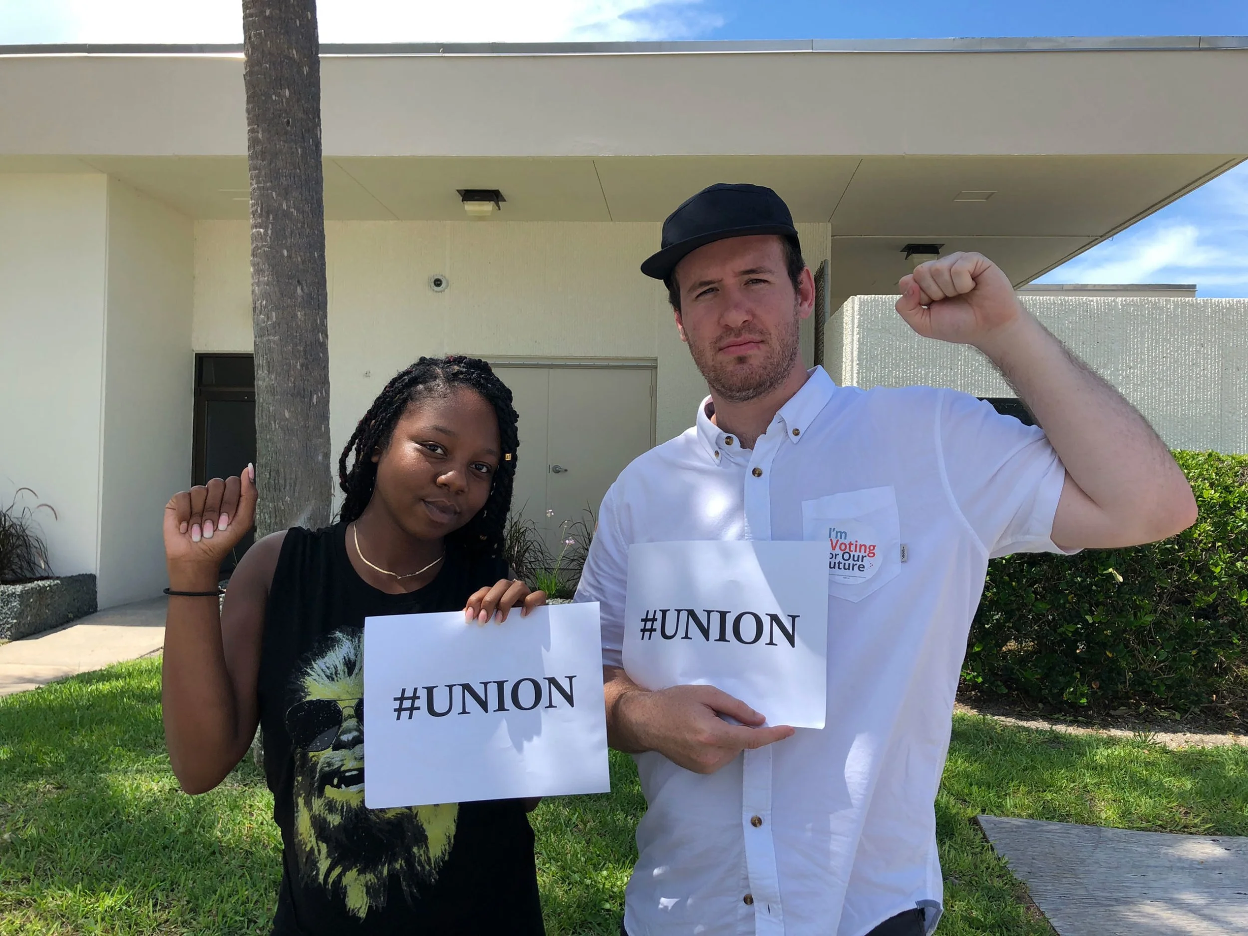 Two young protesters, one female and one male, holding signs with #UNION in front of a modern building. The female wears a black sleeveless top with a lion graphic, has black braided hair, and is raising her right fist. The male wears a white button-up shirt, a black cap, and holds a sign while raising his left fist.