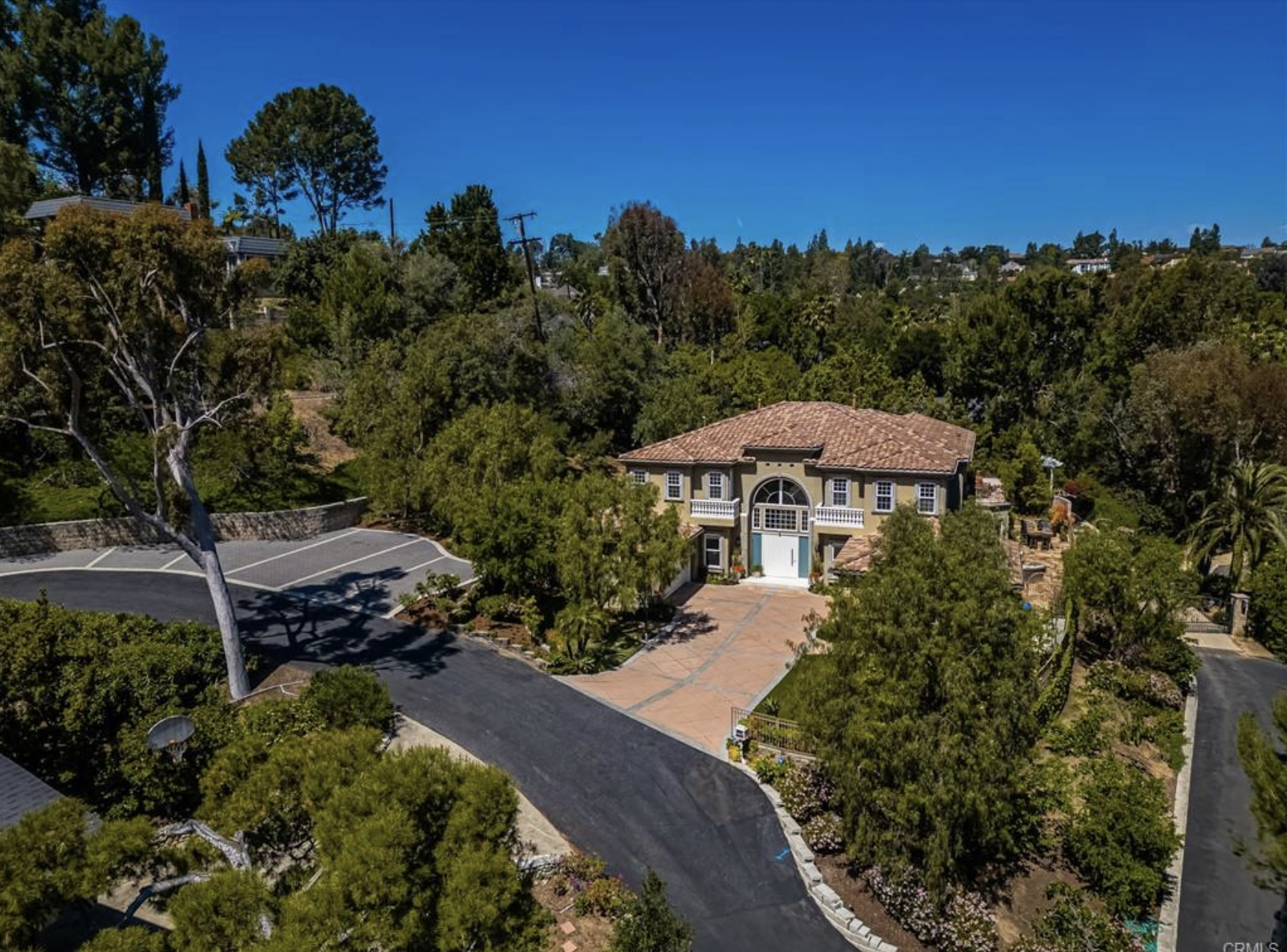 An aerial view of a large house with a tiled roof surrounded by trees and greenery in a hilly area, with a curved driveway and parking lot.