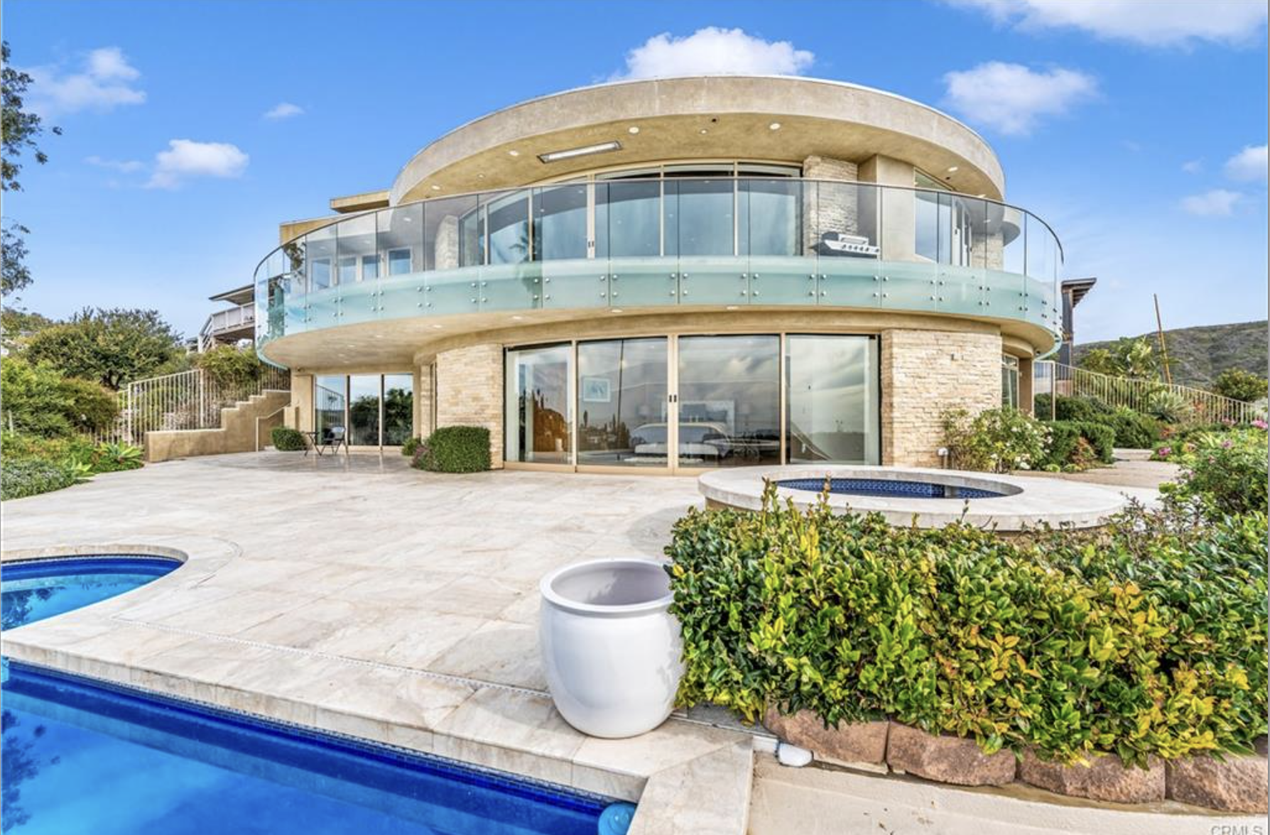 Modern house with large glass windows and balcony, outdoor patio with small pools, surrounded by greenery and garden plants under a blue sky.
