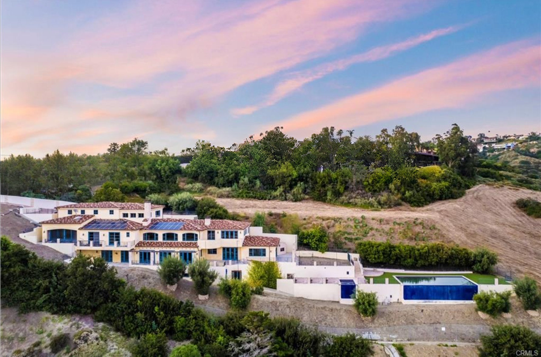 Luxury house with solar panels on a hillside with green trees, a pool, and a sunset sky.