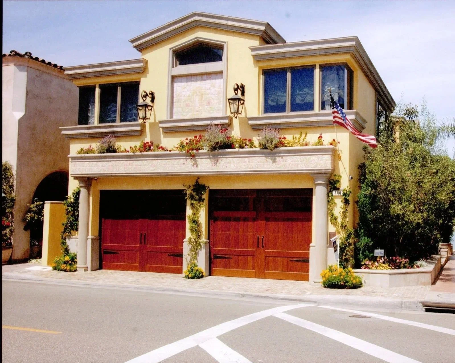 A two-story house with a yellow exterior and a red double garage door. There are large windows on both floors, and the house has decorative columns at the entrance. An American flag is hanging outside, and there are flower beds with colorful flowers and greenery in front and on the balcony. The house is located on a street corner with a sidewalk, and the sky is clear with some clouds.