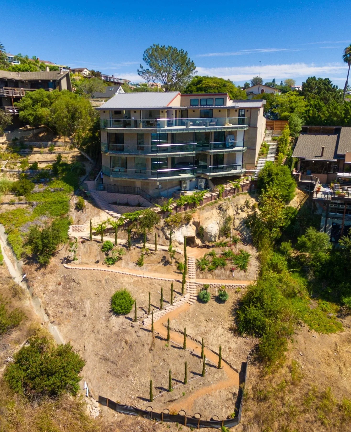 A modern, multi-story house on a hillside with multiple balconies and glass railing, surrounded by trees and other houses under a clear blue sky.