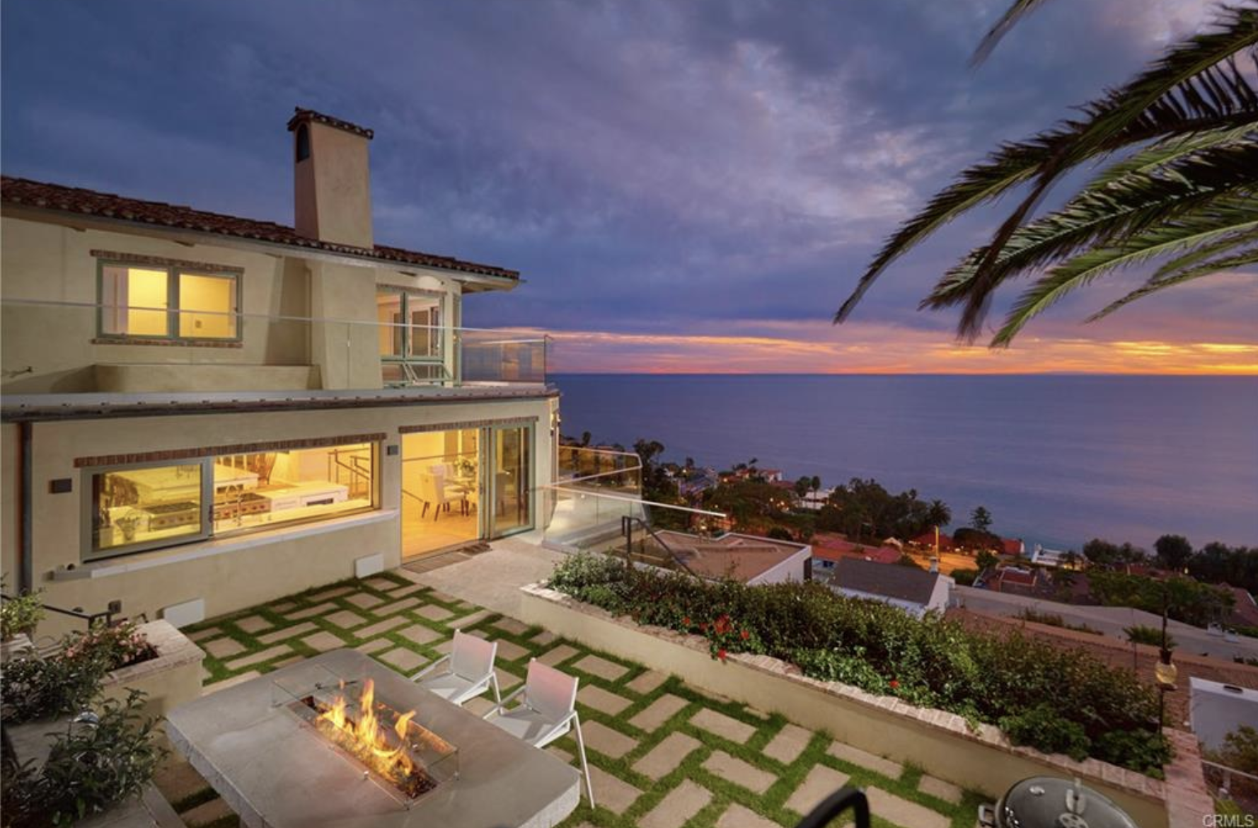 A multi-story house on a hillside overlooks the ocean at sunset, with lit windows, outdoor lighting, and a fire pit on the patio.