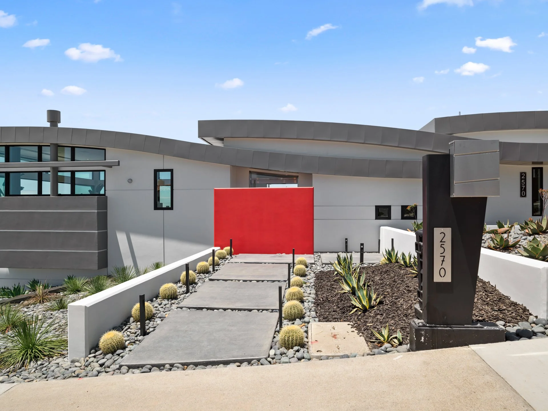 Modern residential house with a minimalist exterior, surrounded by desert plants and cacti, featuring a pathway with large stone slabs leading to the entrance, and a black mailbox with house number 2570.