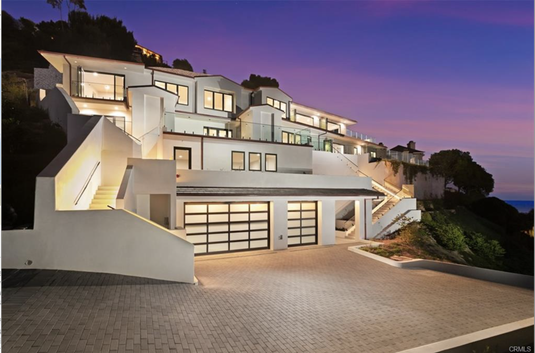 Modern multi-level house on a hillside at dusk with large illuminated windows, outdoor stairs, and a tiled driveway.