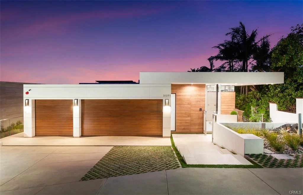 Modern house with three wood garage doors, illuminated exterior lights, and a flat roof, set against a purple and pink sunset sky with palm trees and greenery in the background.