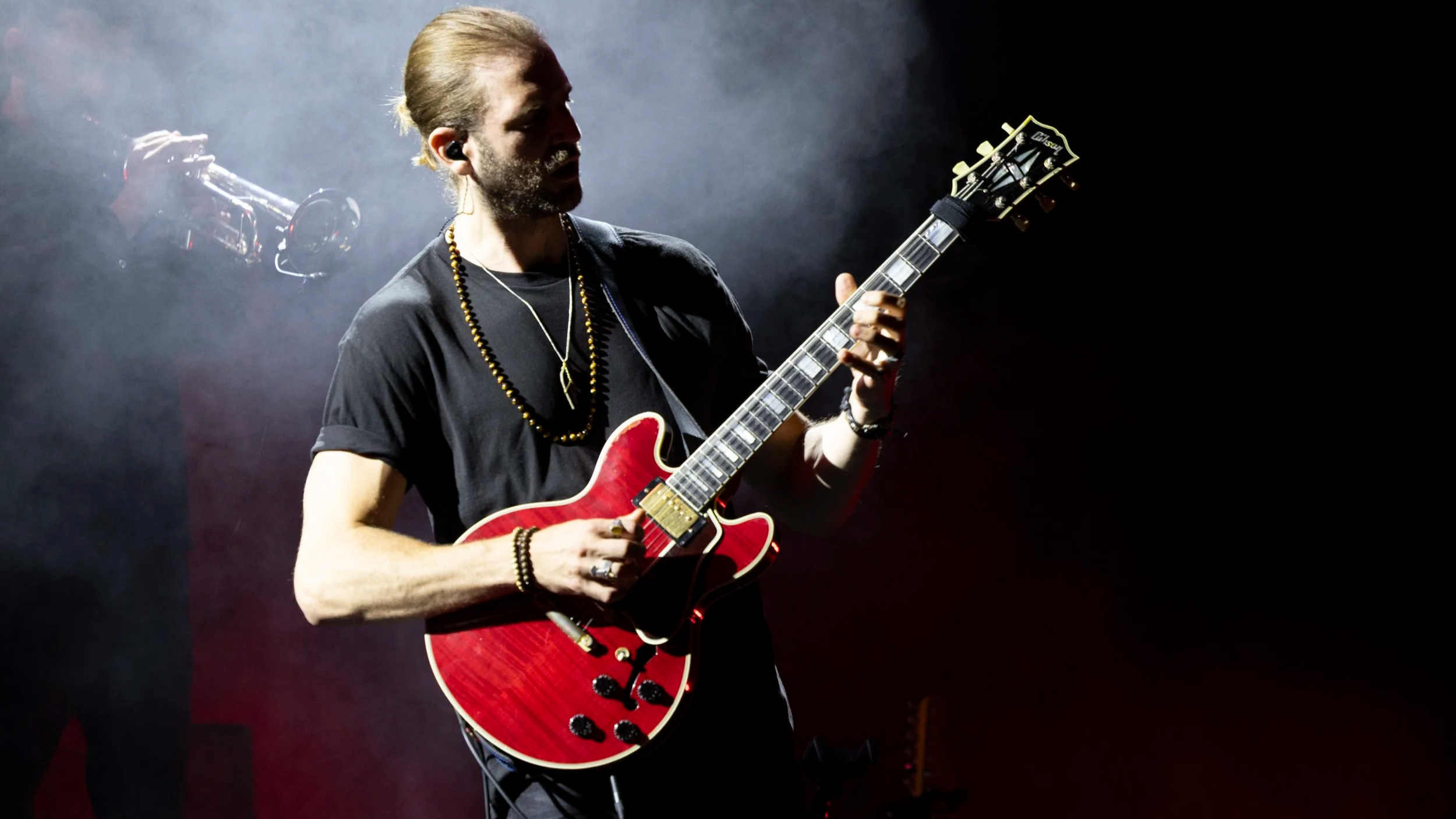 A musician playing a red electric guitar on stage, with a dark background and a trumpet player in the background.