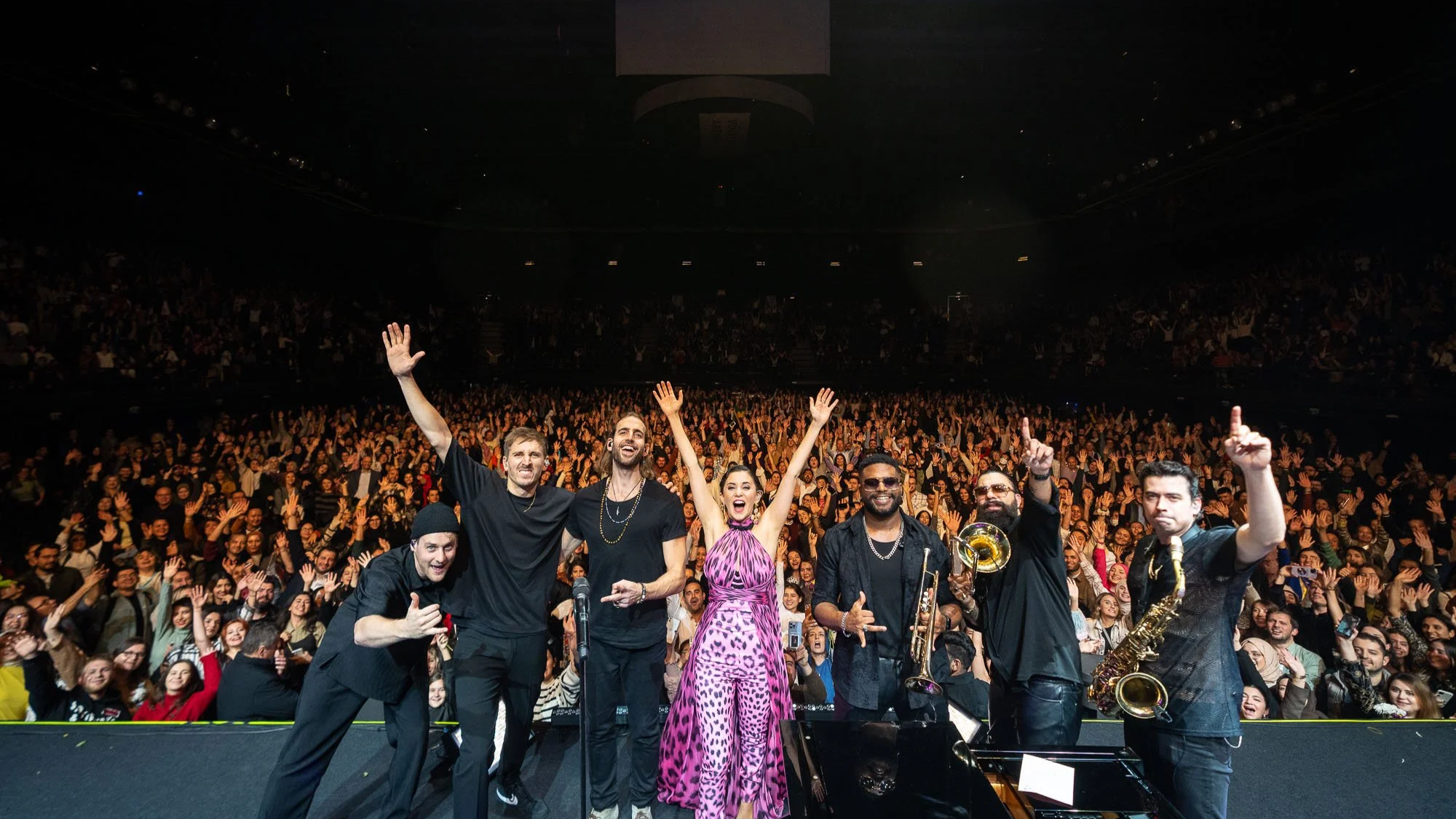 Musical band performing on stage with large audience cheering in the background.