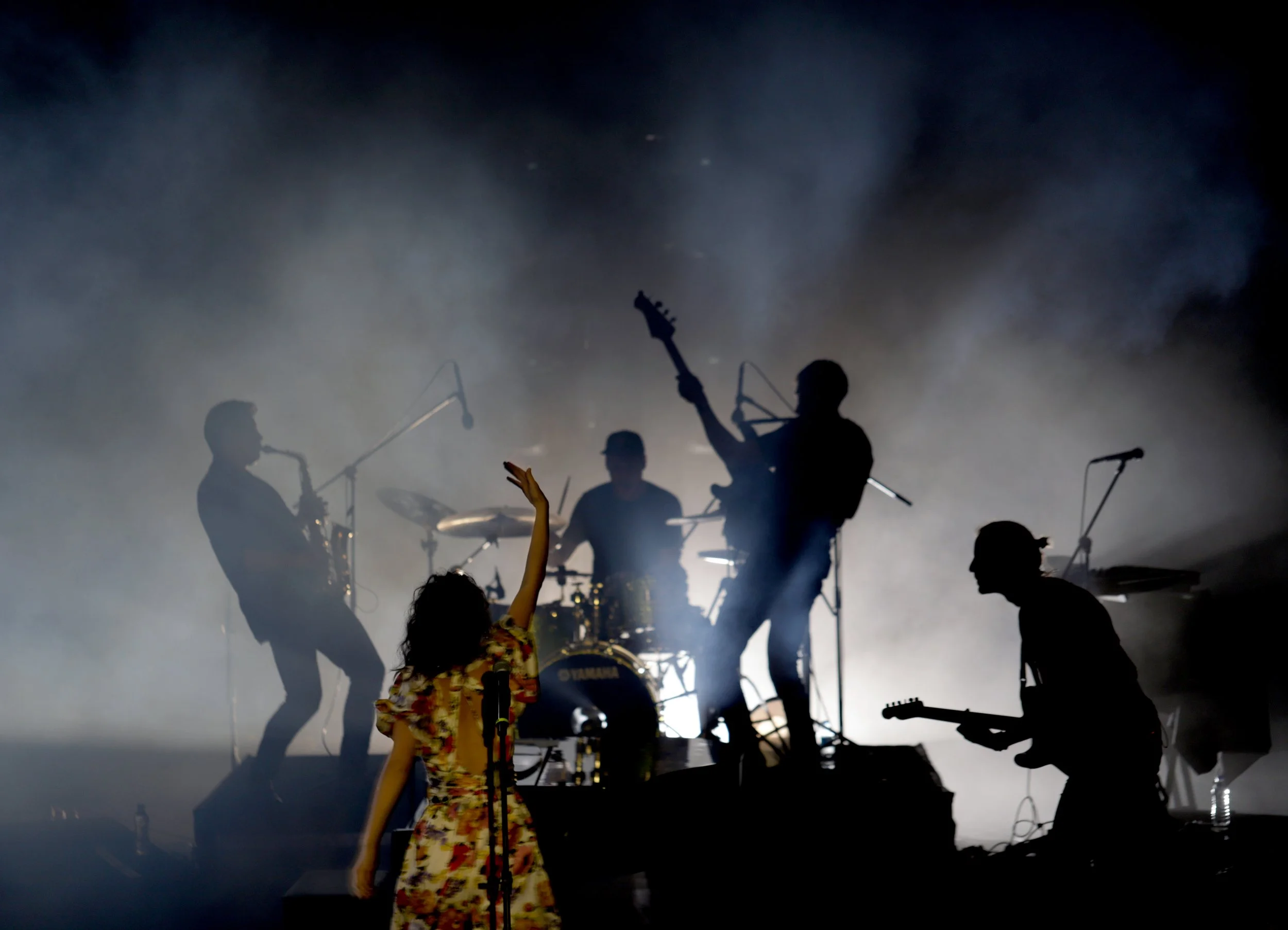 Jessy Hay. Silhouettes of a band performing on stage with a woman singing and raising her hand, illuminated by dramatic backlighting and fog.