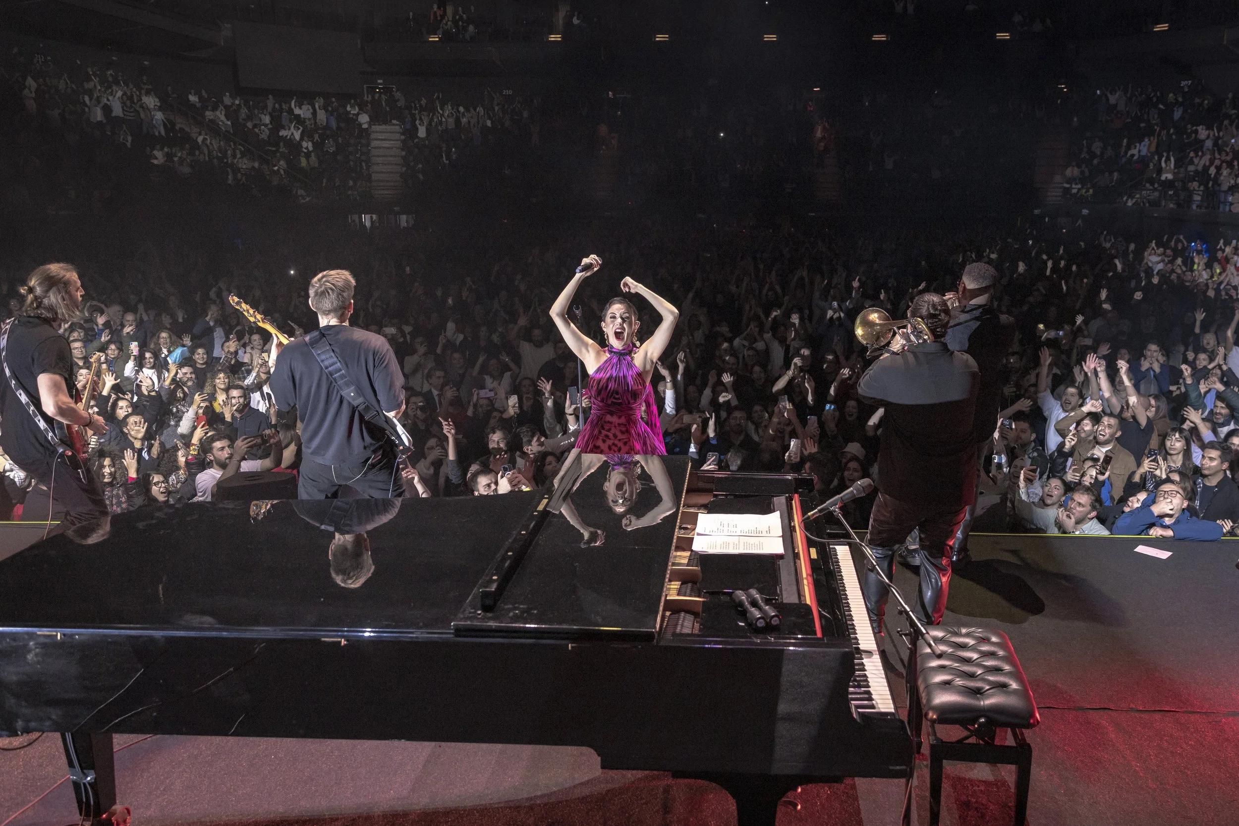 Performer in a pink dress on stage with brass and string musicians, facing a cheering audience in a concert hall.