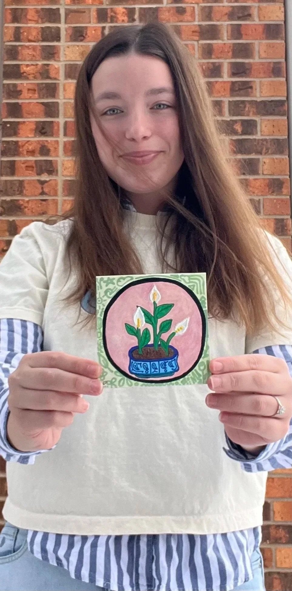 A woman with long brown hair and a light-colored sweater holding a colorful drawing of a potted plant with three candles against a brick wall background.