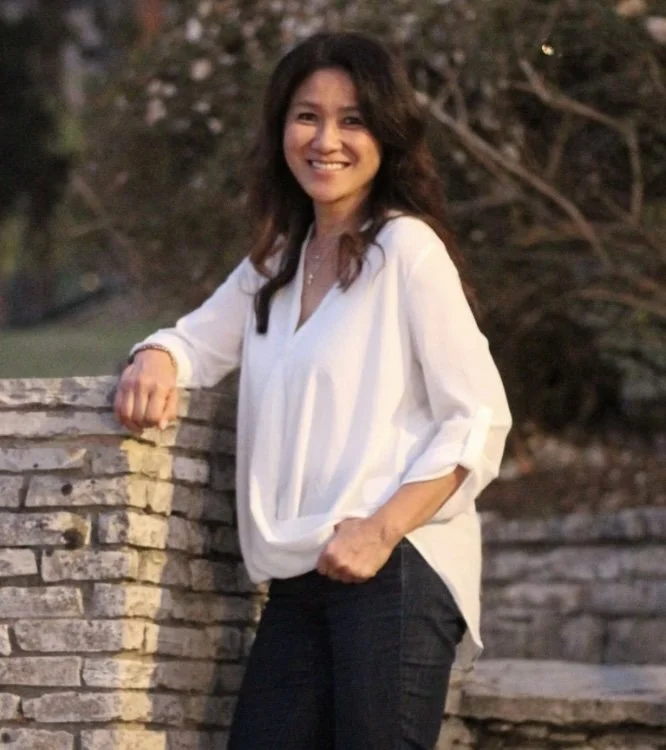 Smiling woman with wavy dark hair wearing a white blouse and dark pants outdoors near a stone wall.
