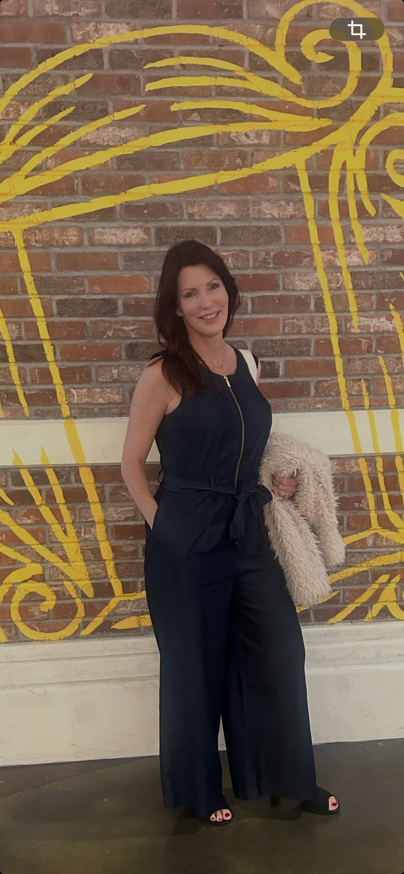 A woman with dark hair wearing a navy sleeveless dress with a zipper and holding a beige coat, standing in front of a brick wall with yellow and pink abstract streaks.