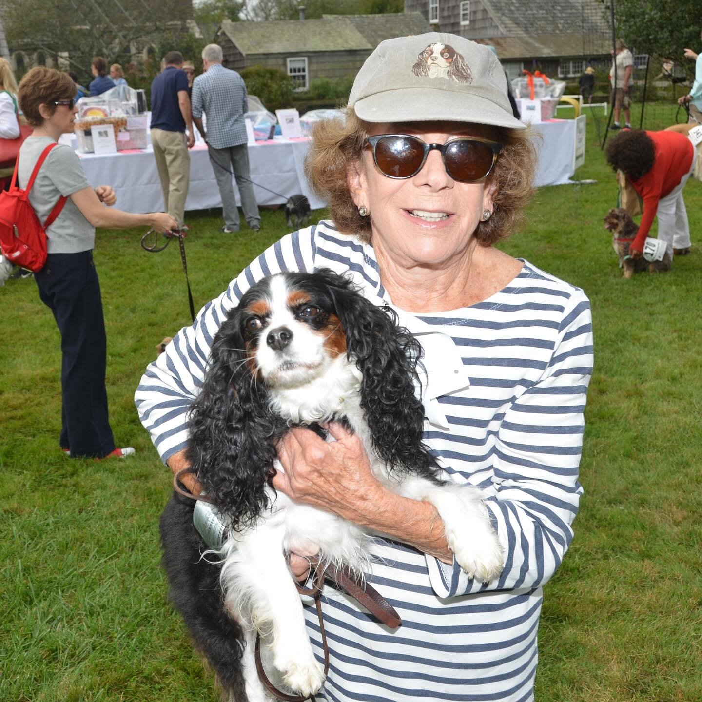 An elderly woman wearing sunglasses, a striped shirt, and a beige hat is holding a black and white Cavalier King Charles Spaniel dog at an outdoor dog event on a grassy field, with people and other dogs in the background.