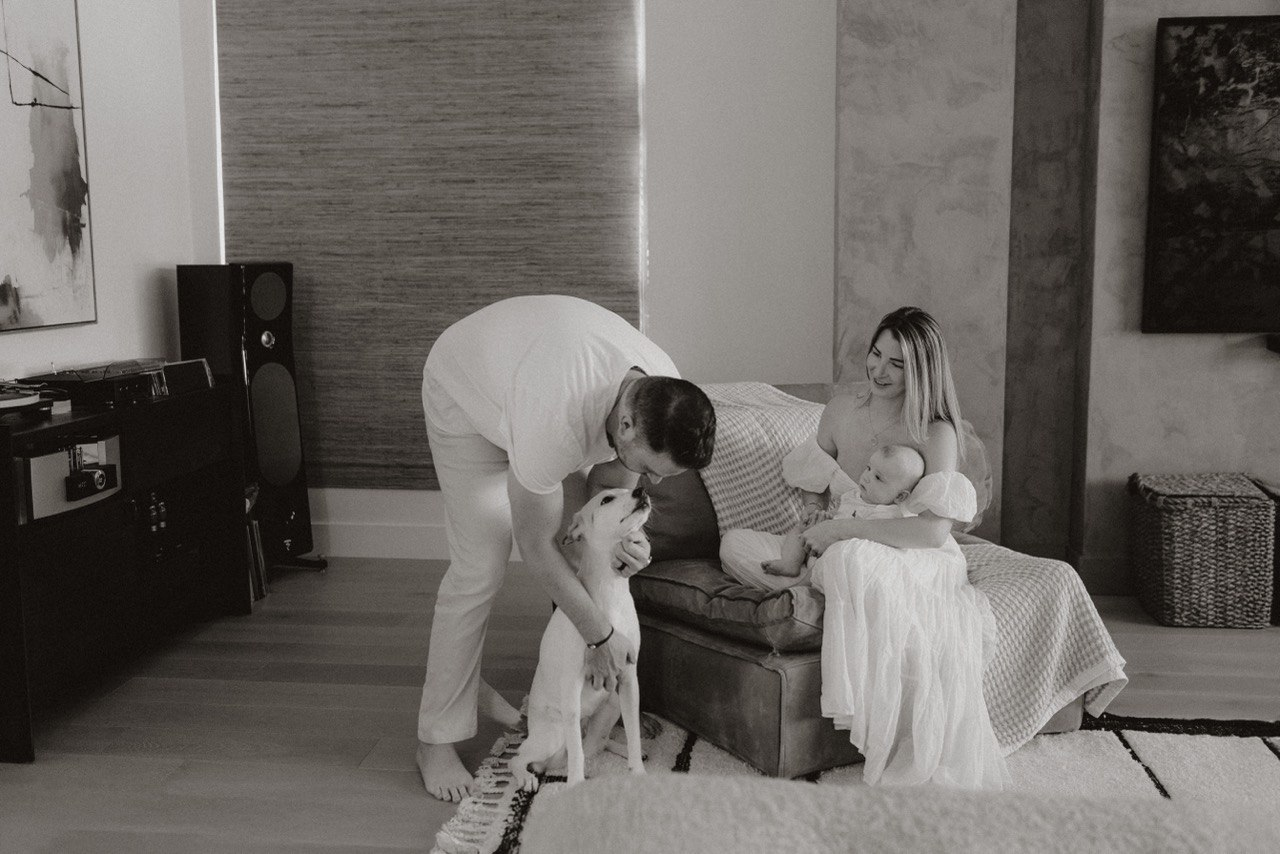 A family scene in a living room showing a woman sitting on a couch holding a baby, a man bending down petting a dog, with a motherly figure watching and smiling.