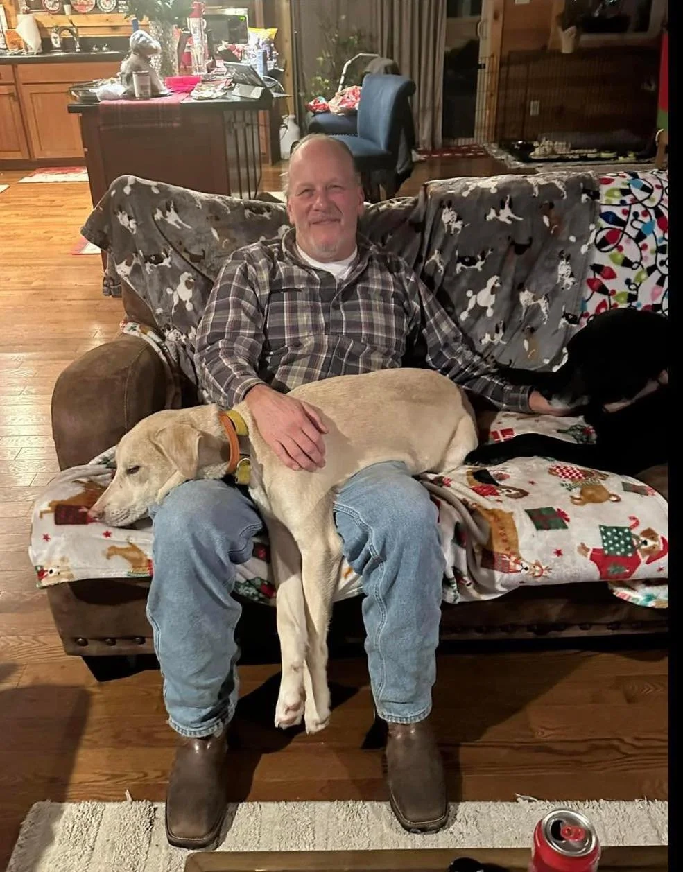 A smiling man in a plaid shirt and jeans sitting on a sofa with two dogs, one tan and one black, in a cozy living room decorated for Christmas.