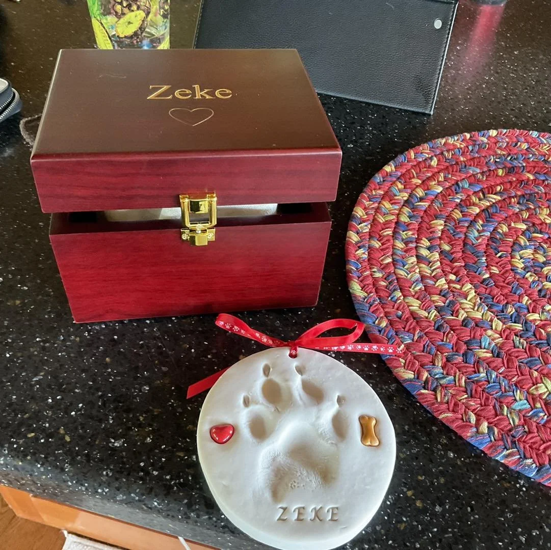 A dark wooden jewelry box with gold latch, a round pet paw print ornament with a red ribbon, small red heart and bone decorations, and a colorful braided placemat on a black countertop.
