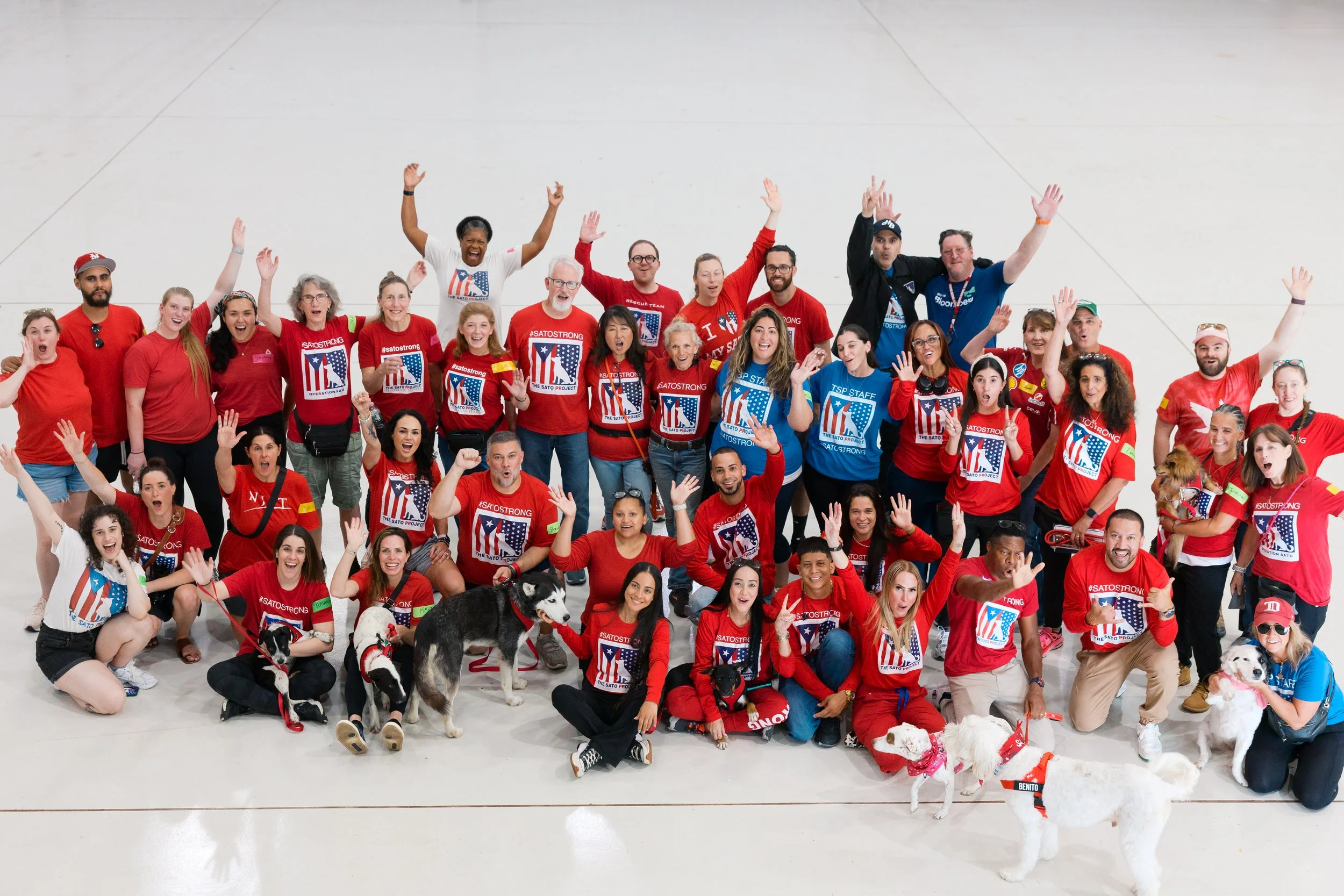 Group of people wearing red and blue shirts, smiling and raising their hands, with dogs and puppies in an open indoor space.