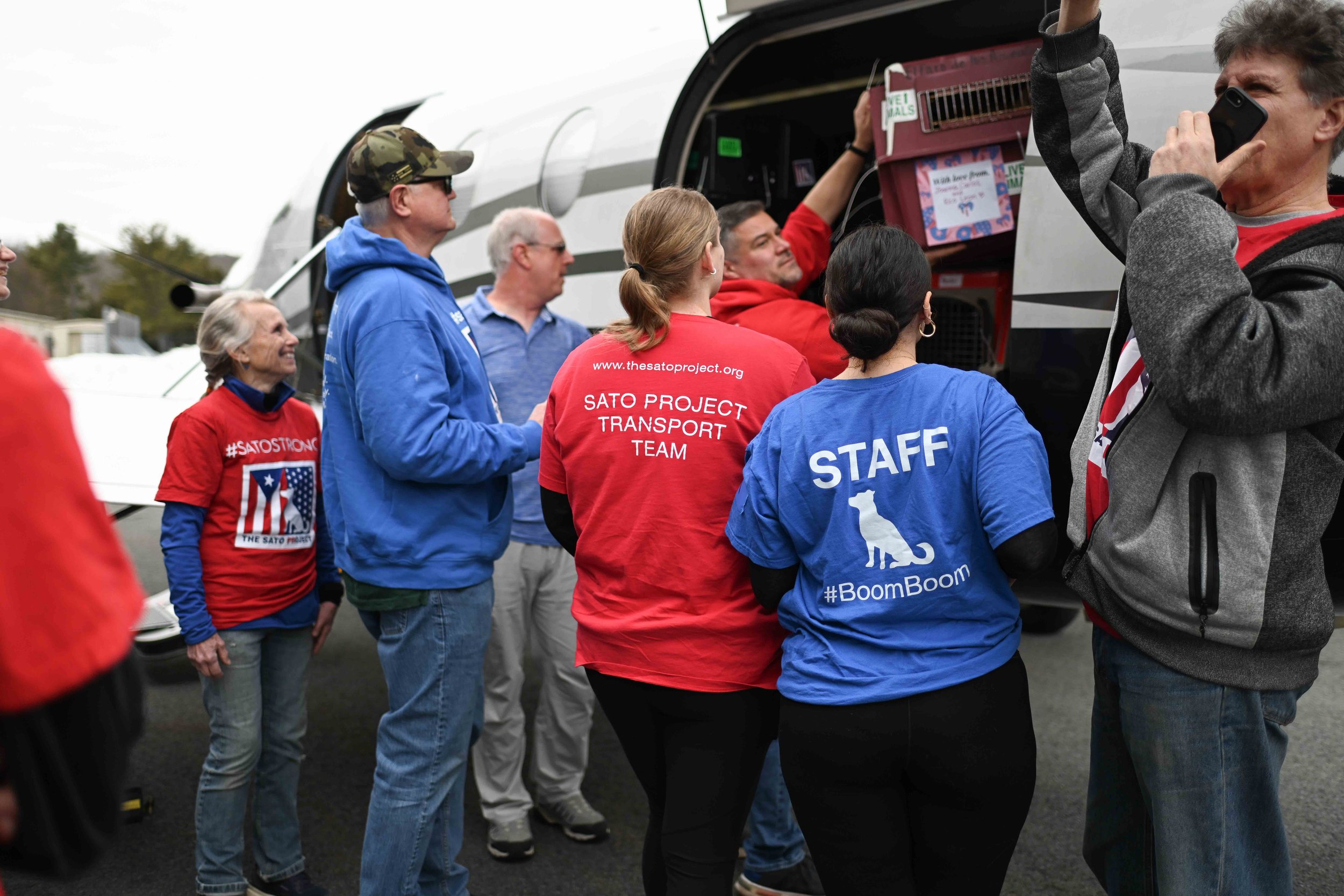 Group of people in red and blue shirts gathering around an aircraft, with some loading luggage into the plane's cargo hold.