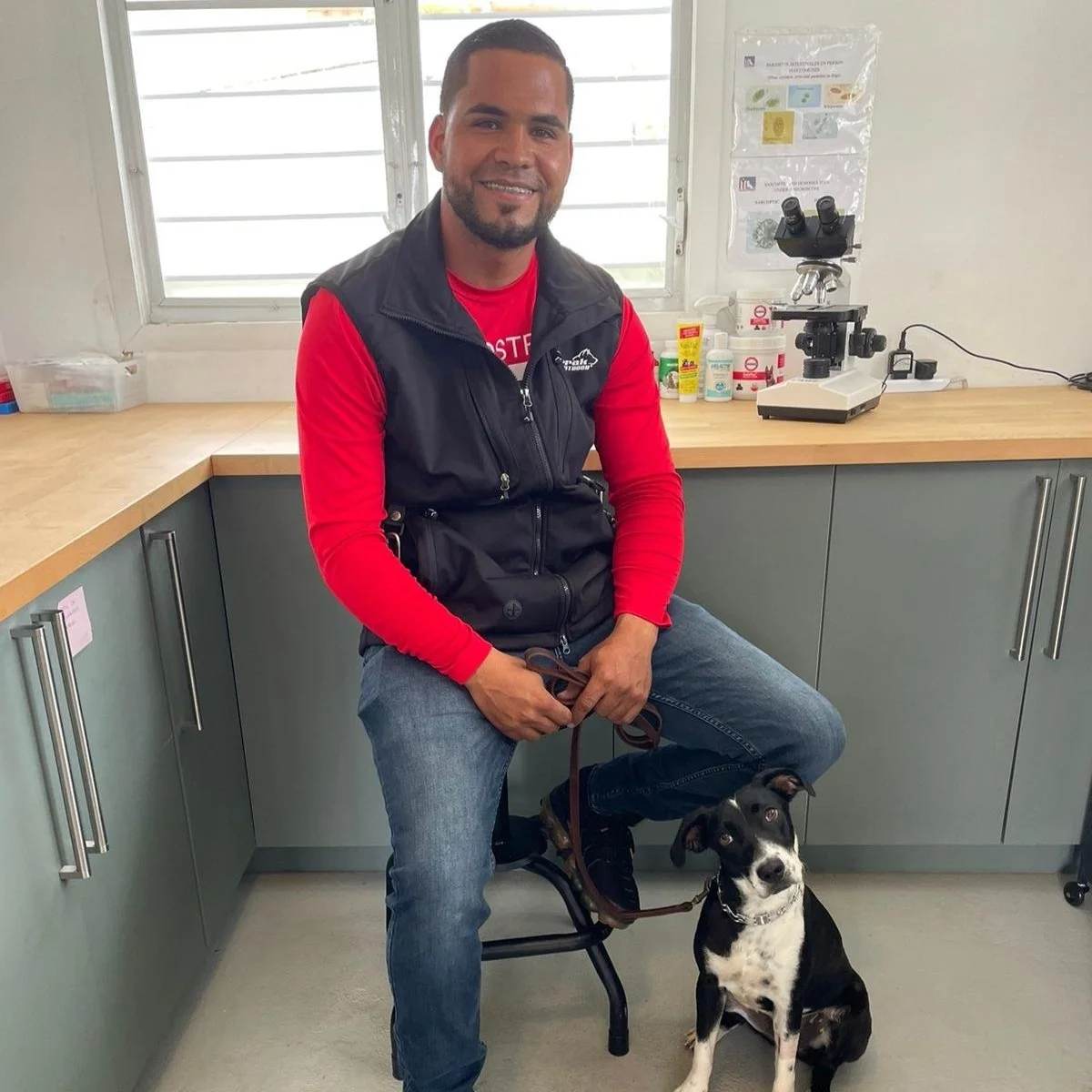 A man sitting on a stool with a black and white dog in a room with a laboratory microscope and supplies on the countertop behind him.