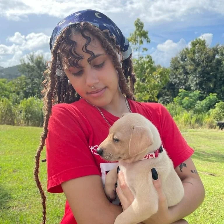 A young woman with curly hair wearing a blue bandana and red shirt holding a small yellow Labrador retriever puppy outdoors on a sunny day with trees and a cloudy sky in the background.