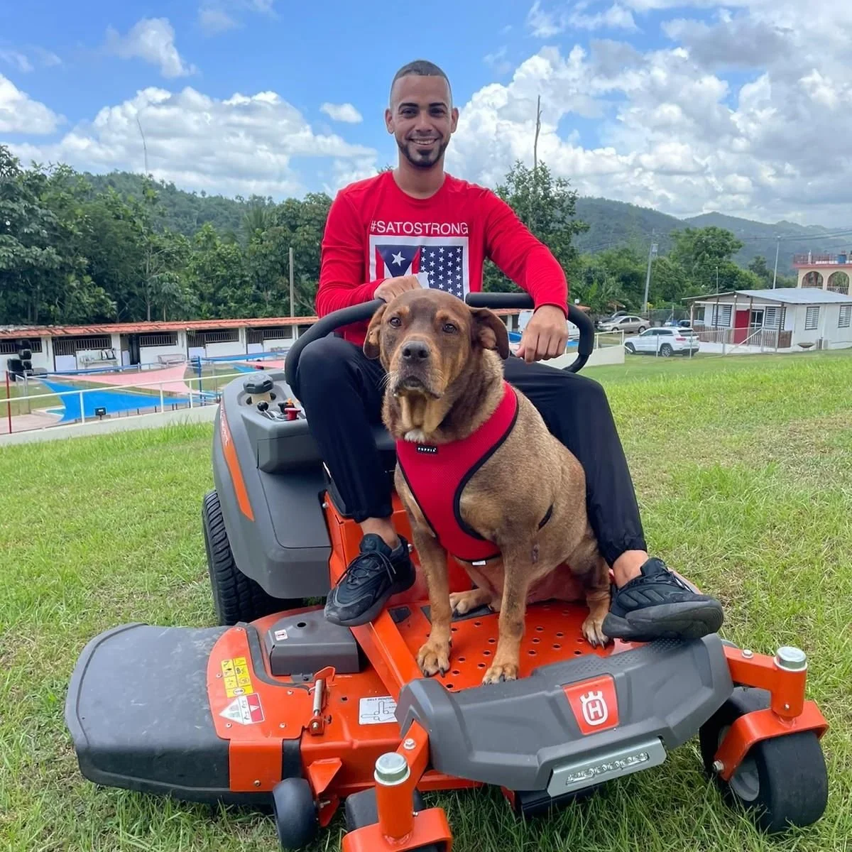 A man sitting on a riding lawn mower with a brown dog sitting on the mower. Both are wearing red clothing, with the man wearing a red shirt and the dog wearing a red vest. The background shows a grassy area, some buildings, trees, and a partly cloudy sky.