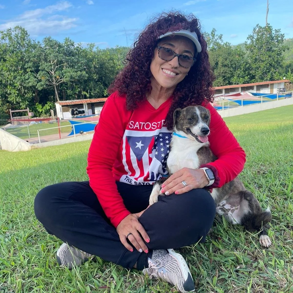 A woman with curly hair wearing glasses, a white visor, and a red long sleeve shirt sitting on grass, holding a small dog with white and brindle fur. They are outdoors with a background of trees, a building, and a sports field.