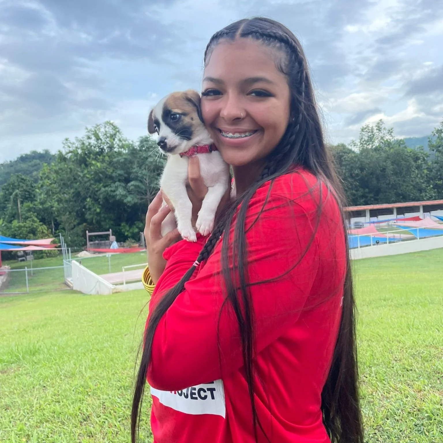 A young woman with long dark hair, wearing a red shirt, smiling and holding a small, white and brown puppy with a pink collar outdoors on a grassy field with trees and a cloudy sky in the background.