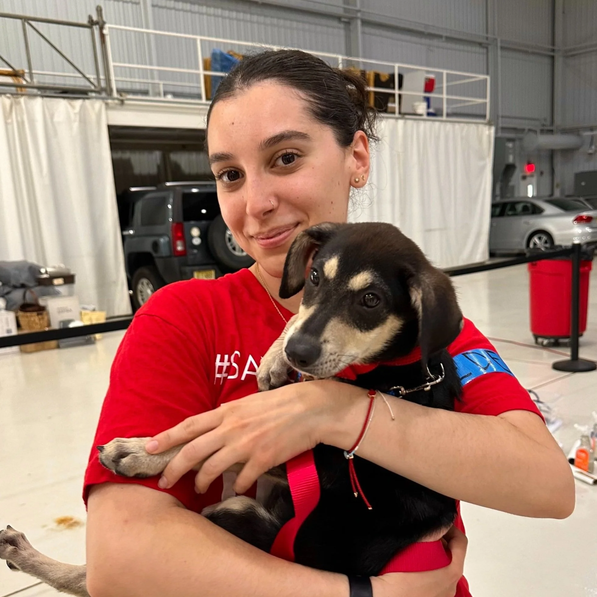 A woman in a red shirt holding a black and tan puppy in an indoor setting, possibly an airport or large facility.
