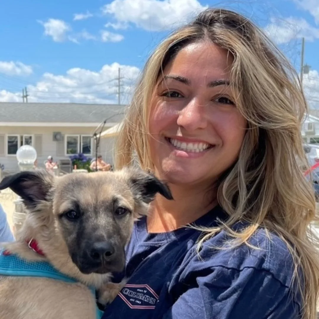 A woman smiling and holding a tan and black puppy outdoors on a sunny day.