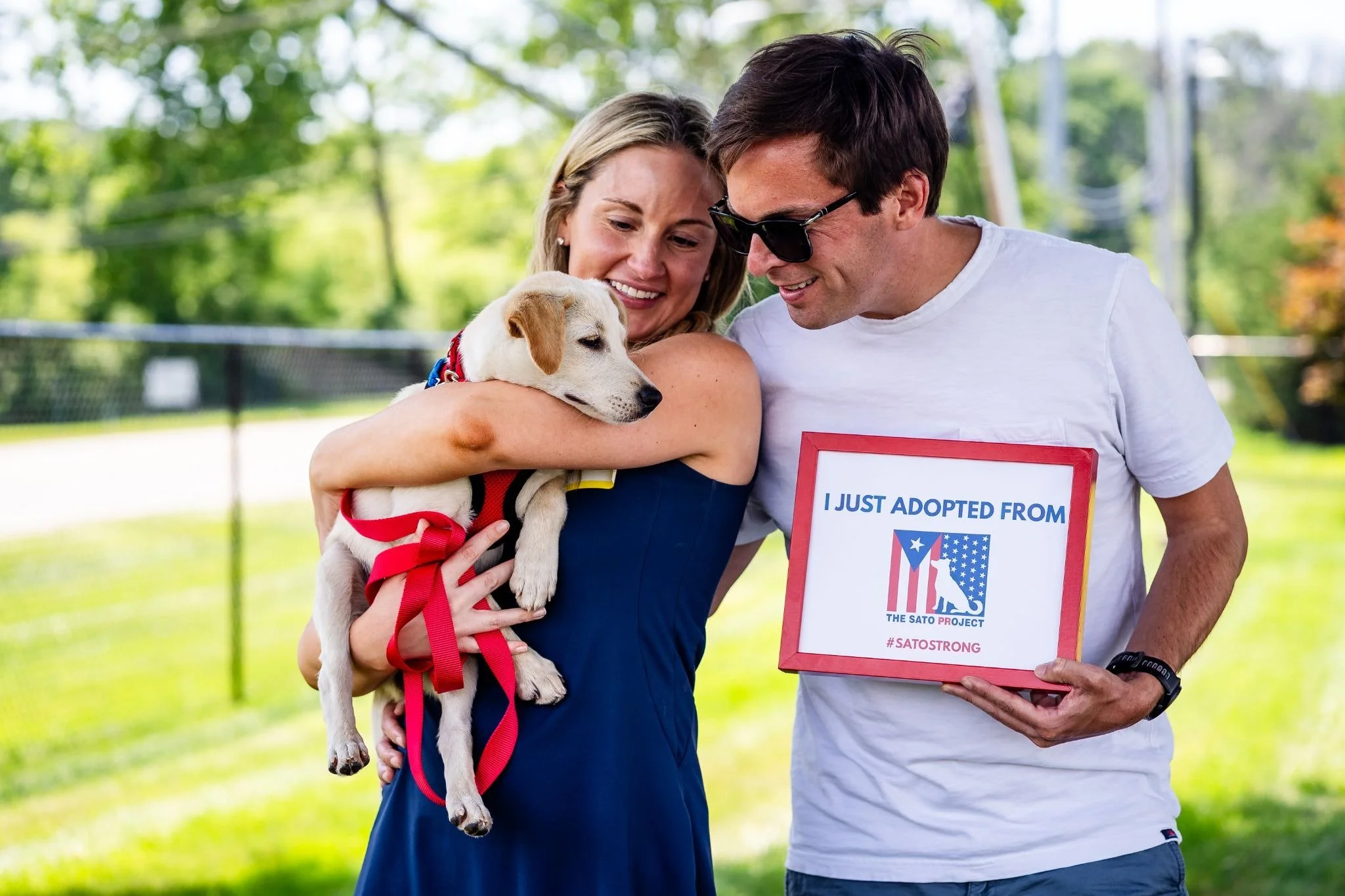 A smiling woman holding a puppy with a red ribbon and a man holding a sign that says 'I just adopted from The SATO Project' outdoors in a park.