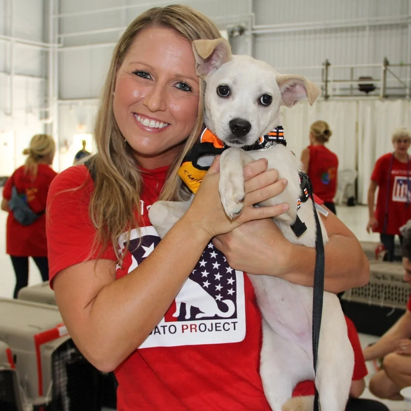 A woman with blonde hair smiling and holding a white puppy with a black nose and dark eyes indoors at an event, with several other people in red shirts in the background.