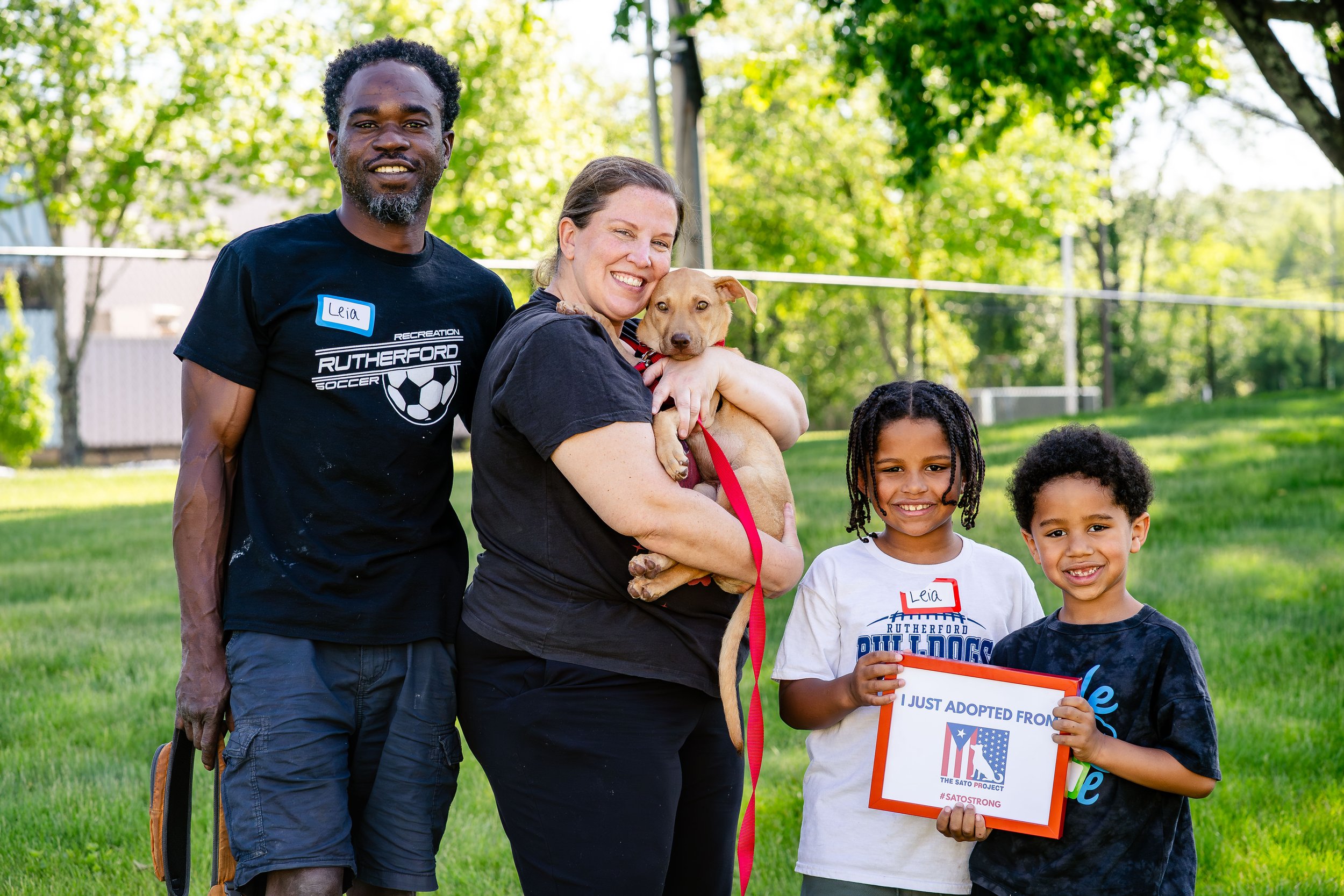 Group of people outdoors, including a man, woman, and children, with one holding a sign indicating they adopted a pet, in a park with green trees.