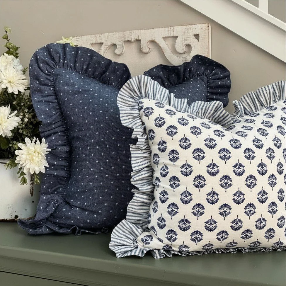Two decorative pillows with ruffled edges, one navy with white polka dots and the other white with blue floral patterns, placed on a green surface next to a white vase with white flowers, with a light-colored decorative panel and a staircase in the background.