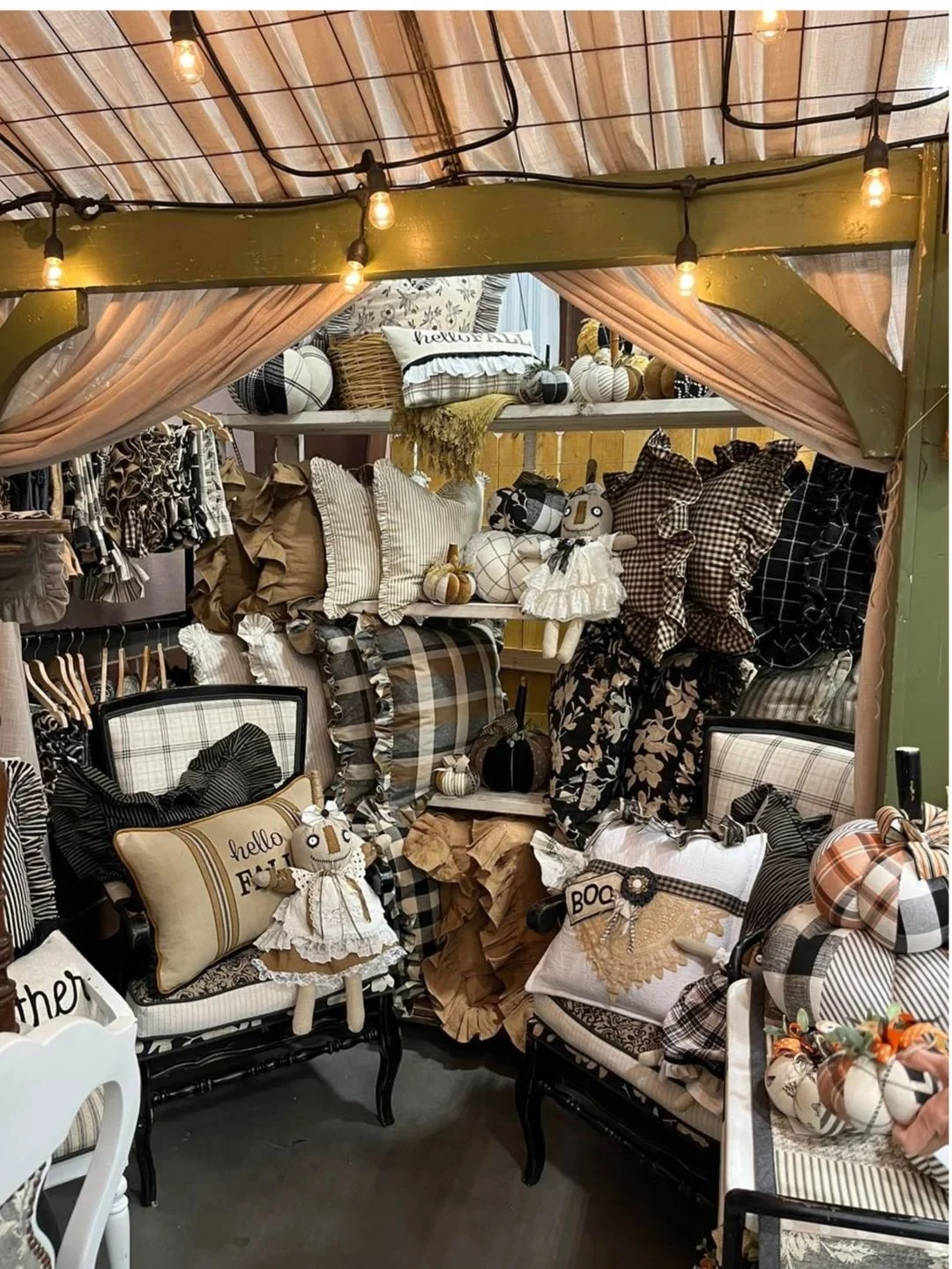 Fall-themed home decor display with plaid and neutral-colored pillows, pumpkins, dolls, and string lights under a canopy with pink curtains.