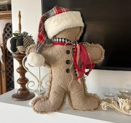 Plush teddy bear wearing a Santa hat and scarf, standing on a white surface with decorative items nearby.