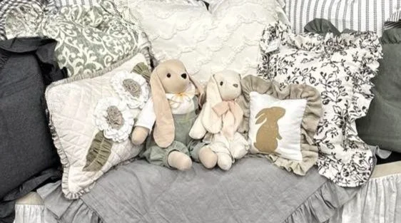 A cozy bed with multiple decorative pillows and two plush bunny toys sitting on the bed, surrounded by a variety of patterned and textured pillows.