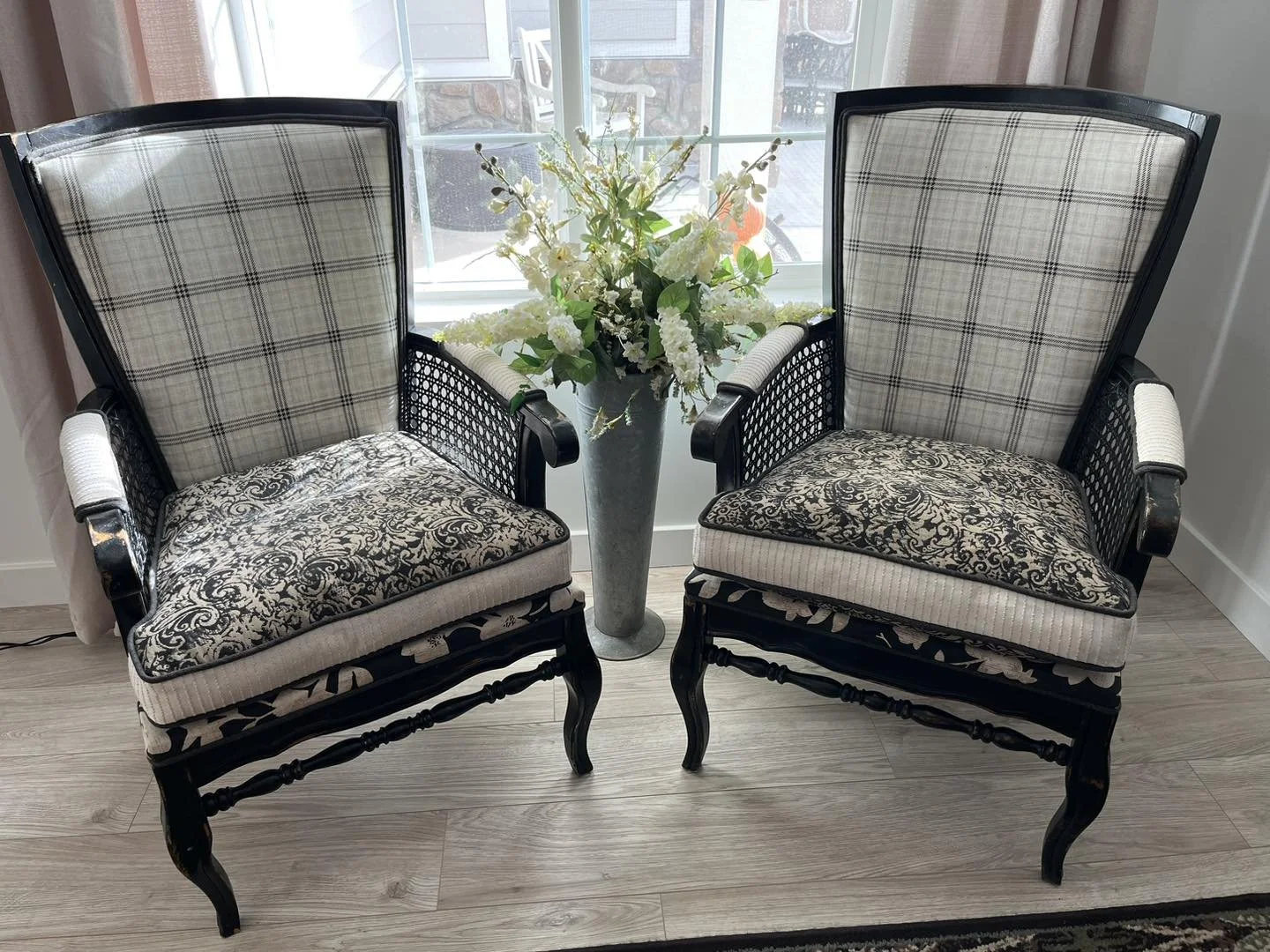 Two vintage black wooden armchairs with patterned cushions and plaid backrests, positioned on a light wood floor, with a tall gray vase holding white and green flowers between them, in front of a glass window with curtains.