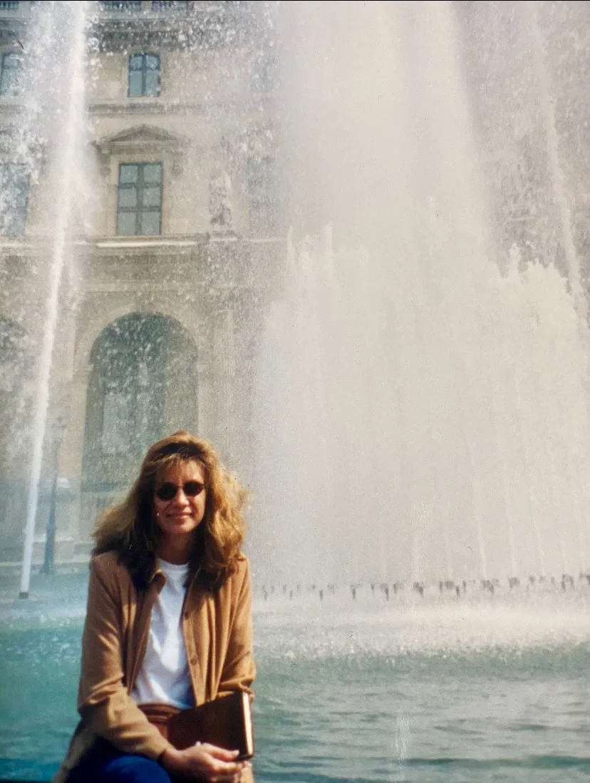 Woman with curly hair, sunglasses, tan jacket, and white shirt sitting in front of a large water fountain.
