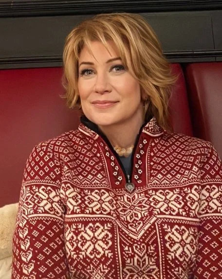 Woman with shoulder-length blonde hair wearing a red and white patterned Christmas sweater, sitting indoors with a red background.