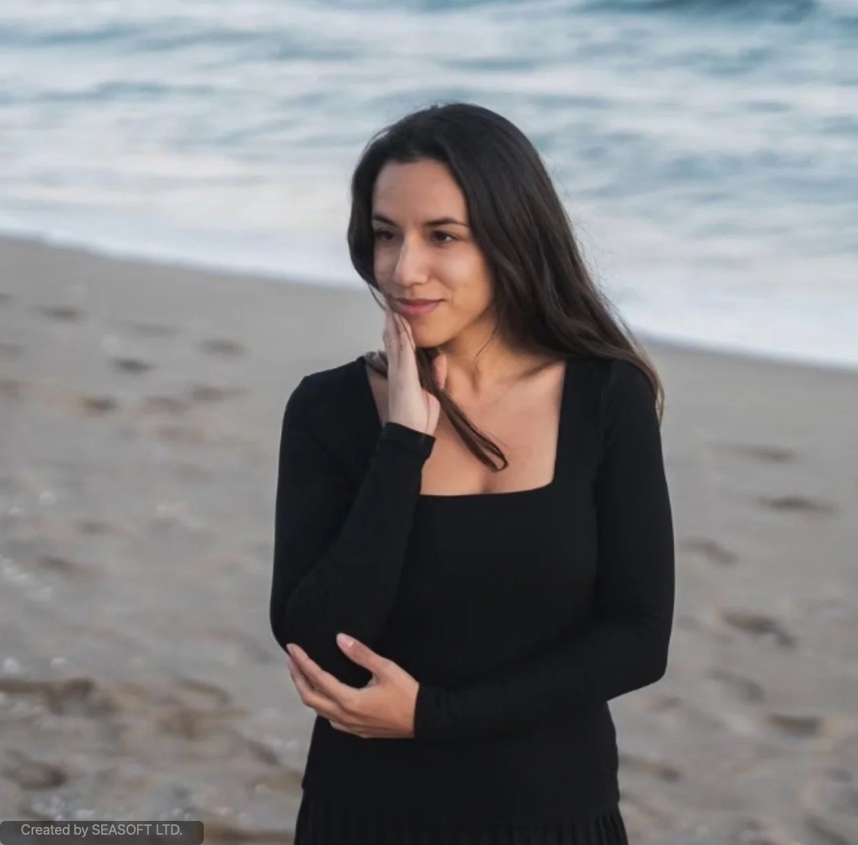 A woman with long dark hair in a black dress is standing on a beach near the ocean, gently touching her face with one hand and holding her other arm. The background shows the shoreline and the water.