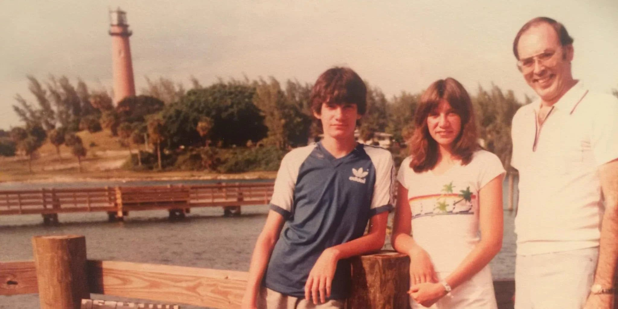 A family of three standing on a wooden dock with a body of water in the background, with a lighthouse, trees, and a hillside visible. The boy is wearing a blue Adidas shirt, the girl is wearing a white t-shirt with colorful palm trees, and the man is wearing glasses and a white polo shirt.