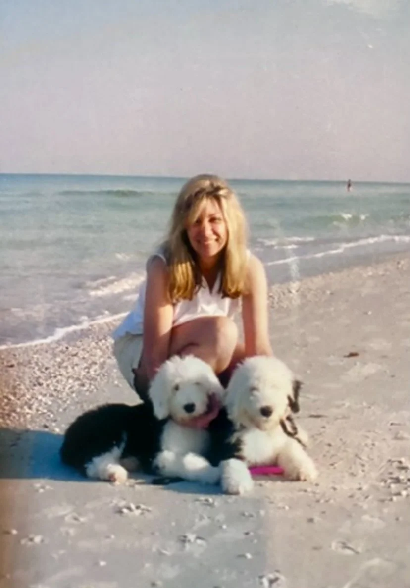 A woman with blonde hair smiling at the camera, crouching on a sandy beach with two black-and-white puppies.