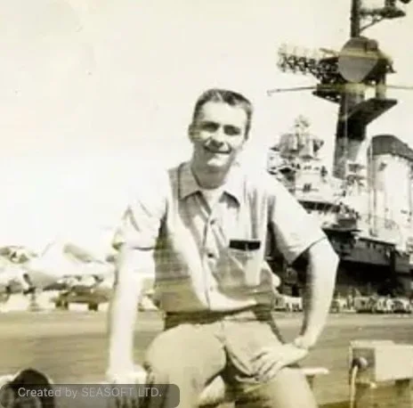 A man in military uniform sitting on a bench at an aircraft carrier, smiling at the camera.